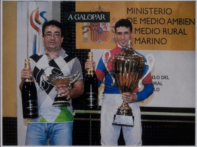 Entrega de trofeos del Gran Premio Costa del Sol: dos hombres sostienen las copas y sendas botellas de champán, uno de ellos con casaca de jinete