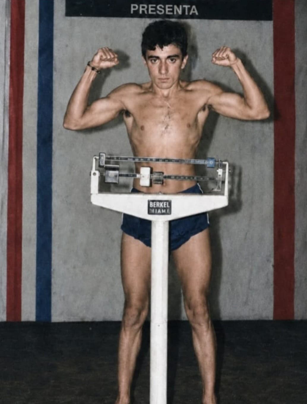 Fernando González Cruz, muy joven, en el pesaje: de pie tras una báscula, sin camiseta y con los dos brazos flexionados, ante una lona de franjas rojas y azules